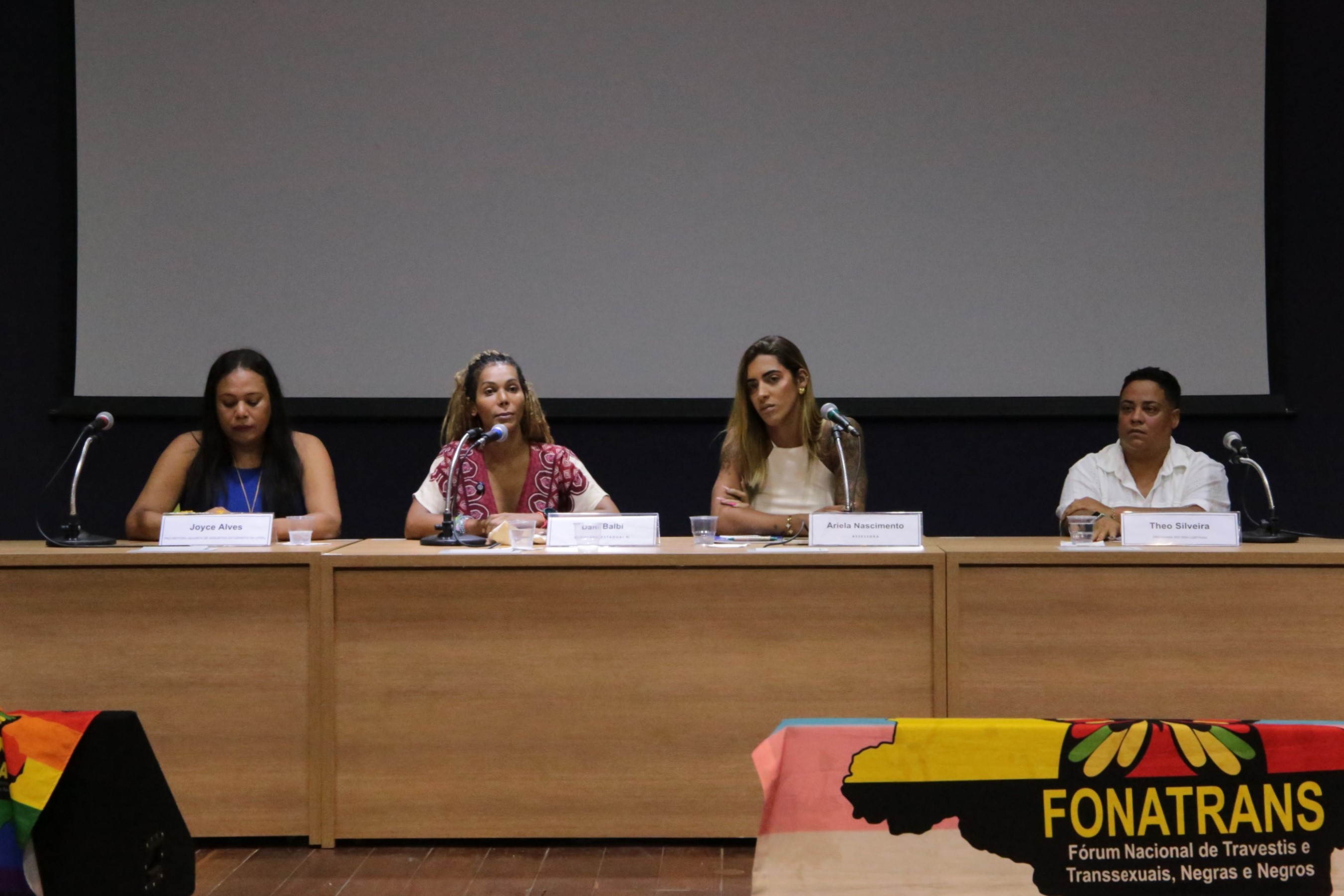 Sentadas em uma mesa comprida com um telão no fundo, da esquerda para direita estão: Joyce Alves, Dani Balbi, Ariela Nascimento e Theo Silveira