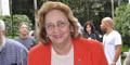 Foto da pesquisadora emérita Dra. Léa Camilo-Couro. Ela veste um blaze vermelho, com uma camisa branca por debaixo do blaze. Ela usa óculos e aparenta ter por volta de 70 anos. Ela está sorrindo.
