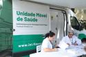 Lateral de uma van, nas cores verde e branco escrito Unidade Móvel de Saúde, Instituto Nacional de Infectologia Evandro Chagas (INI/Fiocruz). Na frente do veículo há duas mulheres sentadas trabalhando. Elas são profissionais de saúde e estão ali para atender as pessoas. Elas vestem roupa branca e ambas aparentam ter cerca de 40 anos. 