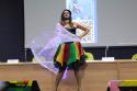 A artista Alessandra Salazary apresenta uma dança usando um vestido colorido de vermelho, amarelo e verde junto com uma capa brilhante.