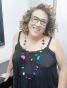Foto de Jacqueline Rocha Cortes. Senhora de meia idade. Ela tem os cabelos na altura dos ombros, na cor castanho claro. Ela usa vestido preto. Ela usa óculos e está sorrindo na foto.  
